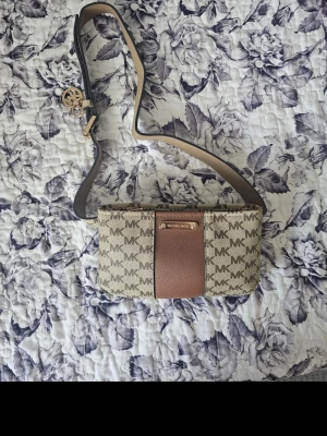Michael Kors magväska med logomönster - Snygg axelväska från Michael Kors i beige och brunt med MK-logga över hela väskan. Väskan har en bred brun rem och en dekorativ metallbricka framtill. Tillverkad i canvas och konstläder med guldfärgade detaljer. Köpt för 1300
