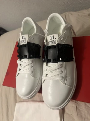HELT NY 😎 Valentino Rockstud Untitled sneakers 41 - Säljer ett par vita Valentino Rockstud Untitled sneakers med svart band och ikoniska nitar över vristen. Skorna har klassisk snörning och är tillverkade i skinn för en lyxig känsla. Perfekta för dig som vill sticka ut med en edgy detalj. SKRIV PÅ VITA lådan OCH PÅSE  FÖLJER MED 😎😎‼️‼️