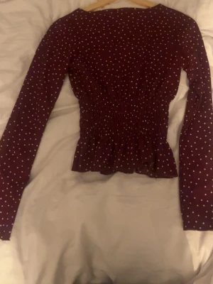 Mörkröd prickig blus med smock - Supersöt mörkröd blus med vita prickar och smockad midja som ger en snygg siluett. Långärmad modell med lätt volang nertill. Perfekt för dig som vill ha en trendig och bekväm topp med lite retrovibbar.