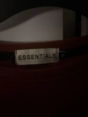 Brun sweatshirt från Essentials - Säljer en brun sweatshirt från Essentials med svart logga tryckt på bröstet. Tröjan har rund halsringning och långa ärmar, perfekt för en avslappnad streetwear-look. 