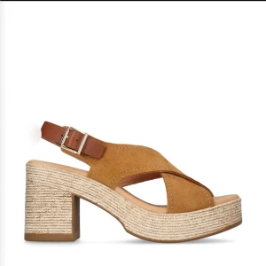 Bruna kilklackade sandaler från Dasia - Snygga sandaler i cognac brun mocka med korslagda remmar och öppen tå. Klack i flätad jute-look och justerbar rem med spänne runt hälen. Perfekta för sommarens alla outfits och ger en avslappnad men trendig vibe. Nypris 1200:-