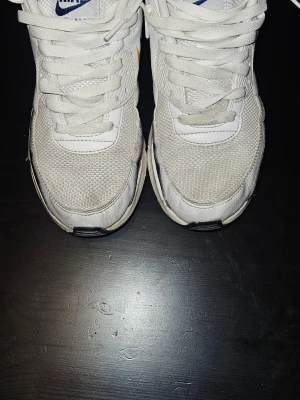 Nike Air Max vita sneakers - Nike Air Max sneakers i vitt med mesh och läderdetaljer. Sidan har en unik Swoosh i gult och blått. Skorna har vita snören och svart sula med synlig Air Max-dämpning. Perfekt för dig som gillar klassisk streetwear med en twist.