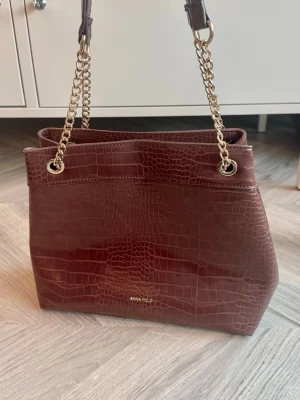 Brun croco handväska från Anna Field - Snygg brun handväska från Anna Field i croco-mönstrat konstläder. Väskan har guldfärgade kedjehandtag och en rymlig fyrkantig form. Perfekt för dig som vill ha en trendig accessoar med lyxig känsla.