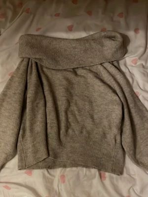 Grå off shoulder stickad tröja - Mysig grå stickad tröja med bred off shoulder-krage och ribbstickade muddar. Tröjan har en avslappnad passform och är perfekt för lager-på-lager-stil. Materialet känns mjukt och stretchigt, vilket gör den extra bekväm.