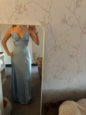 Ljusblå långklänning med slits - Snygg ljusblå långklänning med smala axelband och v-ringad omlottdesign. Klänningen har en hög slits framtill och är tillverkad i ett glansigt material som ger en elegant look. Perfekt för speciella tillfällen såsom bal.