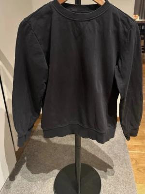 Svart sweatshirt i bomull - En stilren svart sweatshirt med rund halsringning och ribbade muddar vid ärmslut och nederkant. Tröjan har en klassisk passform och är tillverkad i mjukt bomullstyg. Perfekt för en avslappnad look och enkel att matcha med andra plagg. Något urtvättad men i övrigt bra skick