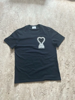 Svart t-shirt från Ami med hjärtlogo - Svart t-shirt från Ami med en stor vit och svart hjärtformad A-logo på bröstet. Klassisk rund hals och korta ärmar. Tillverkad i mjuk bomull, perfekt för en stilren och trendig look.