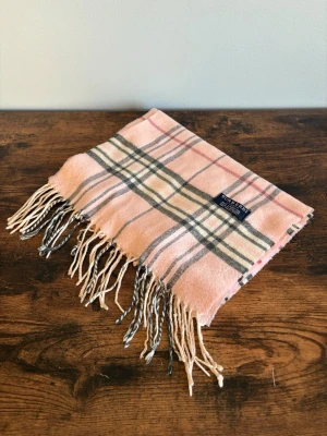 Burberry Halsduk - Burberry halsduk i 100% lambswool. Made in England. Klassisk check i beige/rosa toner med tidlös och stilren look. Mjuk och varm kvalitet med diskret lyxkänsla. Bra skick med mindre tecken på användning. Perfekt för casual/old money stil.