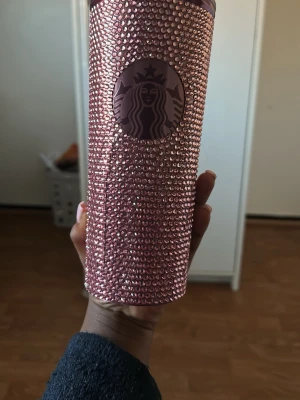 Rosa Starbucks termos med strass - Säljer en snygg rosa termosmugg från Starbucks, täckt med glittrande strass-stenar. Muggen har ett cylinderformat utseende och Starbucks-loggan framtill. Perfekt för att ta med dryck på språng och sticka ut i mängden.