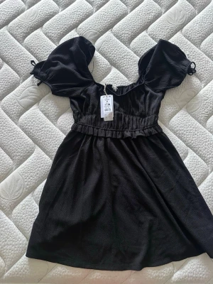 Svart kort puffärmad klänning - Snygg svart kortklänning med puffärmar och söt knytdetalj vid ärmslut. Klänningen har markerad midja och lätt rynkad kjol för en feminin siluett. Perfekt för dig som gillar trendiga och stilrena plagg.