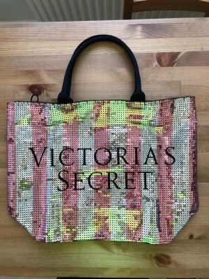 Victoria's Secret paljettväska - Glittrig shoppingväska från Victoria's Secret med paljetter i rosa och silver. Väskan har svarta handtag och stor logga framtill. Perfekt för att bära med sig allt man behöver och sticker verkligen ut med sitt färgglada och glammiga utseende.