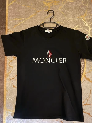 Svart Moncler t-shirt med logga - Svart t-shirt från Moncler med stor vit och röd logga på bröstet samt Moncler-märke på ärmen. Klassisk rund hals och korta ärmar. Perfekt för dig som gillar stilrena och exklusiva plagg.