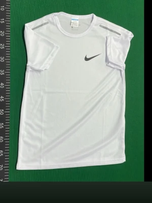 Vit Nike t-shirt i polyester - Sportig vit t-shirt från Nike med klassisk logga på bröstet. Tillverkad i lätt och ventilerande polyester, perfekt för träning. Kortärmad modell med rund halsringning och stilren design.