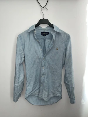 Ljusblå skjorta från Ralph Lauren - Klassisk ljusblå skjorta från Ralph Lauren med lång ärm och knappar framtill. Skjortan har en liten broderad logga på bröstet och är tillverkad i mjuk bomull. Perfekt till jeans eller chinos för en clean look.