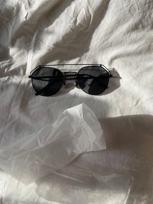 Svarta runda solglasögon i metall - Coola svarta solglasögon med rundade glas och unik dubbel metallbåge. Glasögonen har tunna svarta skalmar och mörka linser som ger en stilren och modern look. Perfekta för att lyfta din outfit med en edgy vibe.