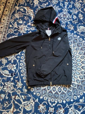 Svart vindjacka från Moncler - Snygg svart vindjacka från Moncler med huva och dragkedja framtill. Jackan har två fickor med tryckknappar, justerbar huva med snören och Moncler-logga på bröstet. Materialet är tunt och lätt, perfekt för blåsiga sommar dagar.