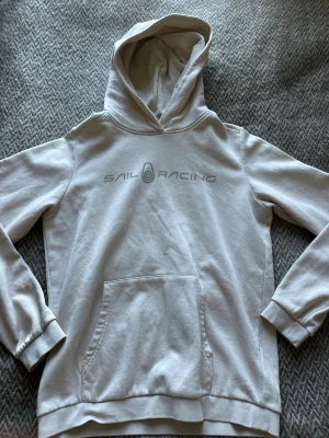 Beige hoodie från Sail Racing - Säljer en stilren beige hoodie från Sail Racing med logga tryckt på bröstet. Hoodien har en klassisk känguruficka framtill, ribbade muddar vid ärmslut och nederkant samt en skön huva. Perfekt för dig som gillar enkel och sportig stil.