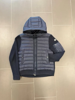 Mörkblå pufferjacka från Moncler - Snygg mörkblå pufferjacka från Moncler med stickade ärmar och huva. Jackan har två fickor med knappar framtill och en ärmficka med dragkedja samt Moncler-logga på vänster ärm. Perfekt för kalla dagar med sin kombination av dun och stickat material.