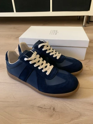 Maison Margiela navy sneakers - Snygga sneakers från Maison Margiela i mörkblå mocka och läder med beige snörning och gummisula. Klassisk low-top modell med stilren design och diskreta detaljer. Perfekta för dig som gillar minimalistisk och trendig stil.