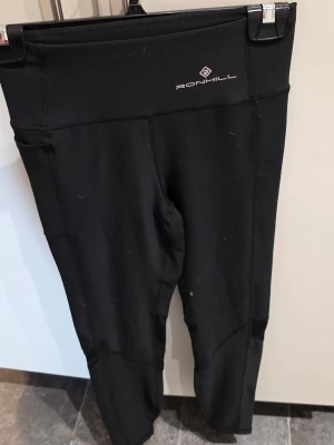 Svarta träningstights från Ronhill - Snygga svarta träningstights från Ronhill med hög midja och diskret logga framtill. Tillverkade i stretchigt funktionsmaterial som passar perfekt för löpning eller gym. Tightsen har sömmar som ger en sportig look och sitter skönt på kroppen.