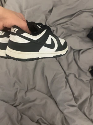 Nike Dunk Low svartvita sneakers - Snygga Nike Dunk Low sneakers i svart och vitt läder med klassisk swoosh-logga på sidorna. Skorna har svart snörning, perforerad tåbox och vit mellansula. Perfekta för dig som gillar streetwear och vill ha en ikonisk look. Har bara använts några gånger 
