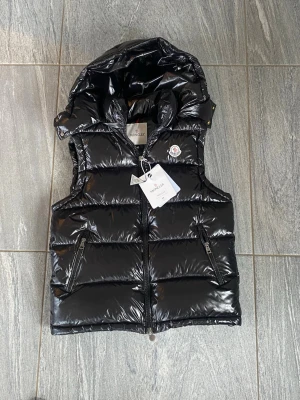 Svart dunväst från Moncler - Snygg svart dunväst från Moncler med glansig finish och avtagbar huva. Västen har två dragkedjeförsedda fickor och klassisk Moncler-logga på bröstet. Perfekt för lager-på-lager och håller dig varm under kyliga dagar. Säljer den för har en annan väst  av tagbar luva och Scaner funkar 