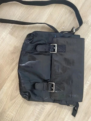 Svart axelväska i skinn från Esprit - Cool svart axelväska i glansigt skinn från Esprit. Väskan har två spännen framtill, justerbar axelrem och lock. Perfekt för dig som vill ha en stilren och rymlig väska med snygga detaljer.