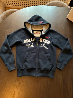 Mörkblå Hollister hoodie med dragkedja - Mörkblå hoodie från Hollister med dragkedja och broderad text 'Hollister Malibu' på bröstet. Har två fickor fram, ribbade muddar och en fodrad huva med grå insida. Klassisk Hollister-logga i rött på bröstet och huvan. Perfekt för en avslappnad stil.