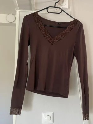 Brun långärmad topp med spets - Mörkbrun långärmad topp med v-ringning och fina spetsdetaljer vid halsringningen och ärmsluten. Toppens material känns mjukt och stretchigt, perfekt för en stilren look. Enkel att matcha med både jeans och kjol.