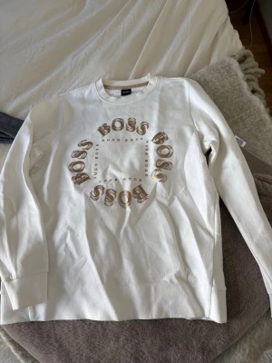 Vit sweatshirt från Hugo Boss - Snygg vit sweatshirt från Hugo Boss med rund halsringning och långa ärmar. Framsidan har ett coolt tryck med BOSS-loggan i cirkel och mindre Hugo Boss-tryck i mitten. Tröjan är i mjukt material och har ribbade muddar vid ärmslut och nederkant. Sitter som en S