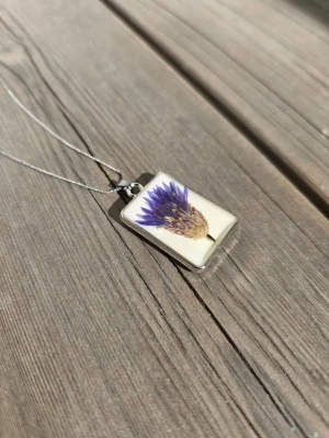 Halsband med blåklint - Unikt halsband med rektangulärt hänge i transparent resin där en torkad blåklimt inbäddad. Hänget har en silverfärgad ram och kedjan är tunn och silverfärgad. Perfekt för dig som gillar naturinspirerade smycken och vill ha något personligt.