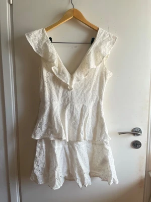 Vit klänning går att ha off shoulder - Studentklänning som man kan ha off shoulder om man vill. Superfin vit byxdress med djup V-ringning och volangdetaljer längs axlarna. Tillverkad i ett broderat bomullstyg med hålmönster och lätt utställda shorts. Perfekt för sommarens alla tillfällen!