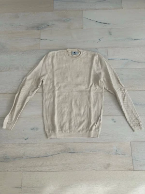 Beige stickad tröja från NN07 - Snygg beige ribbstickad tröja med rund halsringning och långa ärmar. Tröjan har en enkel och stilren design med ribbade muddar vid ärmslut och nederkant. Perfekt för lager-på-lager och passar till det mesta.