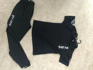 Svart Paris set med tryck - Snyggt svart set med t-shirt och byxor, båda med vita tryck där det står 'PARIS' och emblem på ärmarna. Setet har en sportig look och är gjort i ett mjukt, stretchigt material som känns bekvämt mot huden. Perfekt för dig som gillar streetwear och enkel stil.