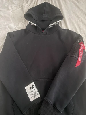 Svart hoodie från Alpha Industries - Snygg svart hoodie från Alpha Industries med vit text på huvan och en ikonisk röd tag på ärmen. Hoodien har en stor magficka och ribbade muddar. Perfekt för dig som gillar streetwear och vill sticka ut lite extra. Pris kan diskuteras.