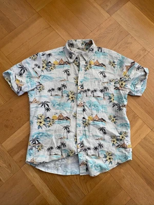 Blommig kortärmad skjorta med palmer - Säljer en beigekortärmad skjorta från Dressmann med tropiskt mönster. Skjortan har knappar framtill, klassisk krage och en bröstficka. Perfekt för dig som gillar avslappnad stil och sommarkänsla.