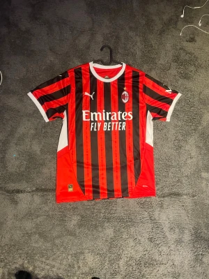 AC Milan matchtröja  röd/svart - Säljer en AC Milan matchtröja i rött och svart med vita detaljer. Tröjan har klubbmärke och sponsortryck på bröstet samt klassiska vertikala ränder. Perfekt för fotbollsfans som vill visa sitt stöd på läktaren eller planen. Passar som M