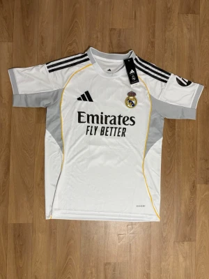 Real Madrid matchtröja Bellingham - Snygg vit Real Madrid matchtröja från Adidas med svarta detaljer och gula linjer. På bröstet finns klubbmärket och sponsorlogga, och på ryggen står det Bellingham samt nummer 5. Tröjan har korta ärmar och är tillverkad i ett lätt, ventilerande material.