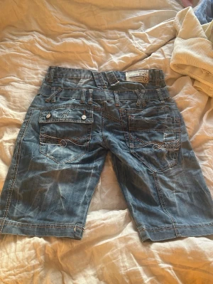 Vintage jeansshorts från Crosshatch - Säljer ett par blå jeansshorts från Crosshatch med vita kontrastsömmar och flera fickor både fram och bak. Shortsen har bälteshällor, knappdetaljer och en avslappnad passform som passar perfekt till sommaren.