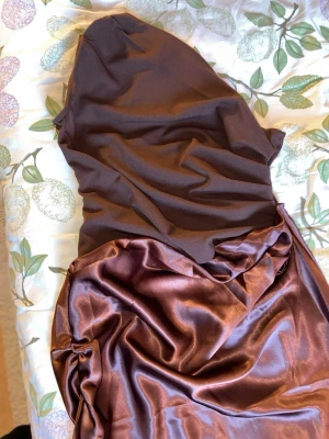 NY! Brun satinklänning  - Snygg lång klänning i två olika bruna nyanser. Överdelen är i matt tyg medan nederdelen är i glansigt satinmaterial med draperad effekt. Ärmlös och figurnära passform. Ny, aldrig använt! 