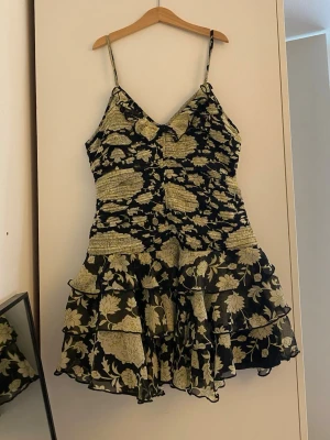 Svart blommig kortklänning med volang - Supersöt kort klänning med tunna axelband och volangig kjol. Svart botten med ljusa blommor i beige och gult. Smickrande v-ringning och omlottkänsla upptill. Tunt, lätt material som känns luftigt och perfekt för varma dagar.
