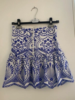 Vit och blå mönstrad kjol från Zara - Supersnygg kort kjol från Zara i vitt och blått med broderat blommönster och dekorativa knappar framtill. Kjolen har hög midja och volangkant nedtill som ger en trendig och somrig vibe. Perfekt för dig som vill sticka ut!