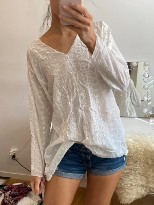 Vit broderad blus med v-ringning - Säljer en luftig vit blus med v-ringning och diskret broderat blommönster. Blusen har lång ärm och är tillverkad i ett tunt, lätt tyg som ger en avslappnad och bohemisk känsla. Perfekt att styla med jeans eller kjol för en fräsch look.