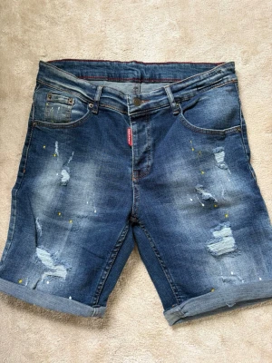 Blå jeansshorts från Dsquared2 - Snygga blå jeansshorts från Dsquared2 med slitna detaljer, färgstänk och uppvikta benslut. Klassisk femficksmodell med knappgylf och normal passform. Perfekta för en avslappnad stil under varma dagar.