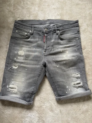 Grå slitna jeansshorts från Dsquared2 - Säljer ett par grå jeansshorts från Dsquared2 med slitna detaljer, färgstänk och upprullade benslut. Klassisk femficksmodell med knappgylf och patch med logga bak i midjan. Perfekta för en avslappnad streetwear-look.
