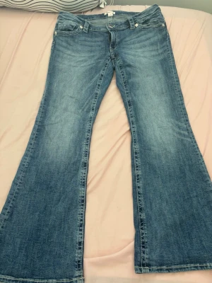 Blå bootcut jeans med utsvängda ben - Säljer ett par klassiska blå bootcut jeans med utsvängda ben och femficksmodell. Jeansen har en ljus tvätt och tydliga slitningar för en cool vintagekänsla. Tillverkade i denim med dragkedja och knapp framtill. Perfekta för en avslappnad och trendig look.