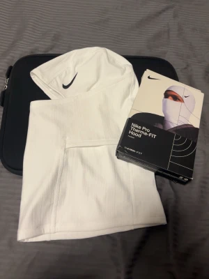 Nike Pro Therma-FIT vit balaclava - Nike Pro Therma-FIT balaclava i vitt, perfekt för kalla dagar. Tillverkad i mjukt och stretchigt material med svart Nike-logga på sidan. Snygg och funktionell huva som täcker huvud och hals.
