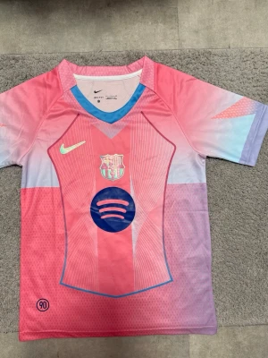 FC Barcelona rosa matchtröja Nike - Säljer en FC Barcelona matchtröja från Nike i rosa och ljusblått med klubbmärke och Spotify-logga på bröstet. Tröjan har korta ärmar, sportig passform och är tillverkad i lätt funktionsmaterial. Perfekt för fotbollsträning eller att bära som supporter.