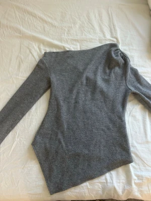 Grå tröja med asymmetrisk kant - Grå tröja i mjukt material med lång ärm och cool asymmetrisk nederkant. Tröjan har rynkade detaljer vid axeln som ger en unik look. Perfekt för dig som gillar stilrena och moderna plagg. Använd ett fåtal gånger. 