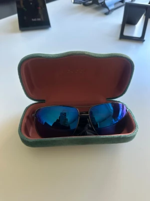 Gucci solglasögon med blå spegelglas - Snygga solglasögon från Gucci med blå spegelglas och metallbågar. Kommer med ett grönt fodral i sammet och röd insida. Glasögonen har en modern, rektangulär form och ger en cool vibe till din stil.